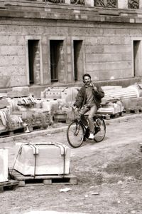 Ein Mann, der mit einem Fahrrad durch eine Baustelle mit gestapelten Steinquadern fährt. Mit einer Hand geschultert trägt er einen Spaten. Im Hintergrund ein historisches Gebäude mit Stuckverzierter Fassade.