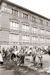 Menschen graben mit Spaten auf einem offenen Gelände vor einem historischen Gebäude mit stuckverzierter Fassade. Eine Seite des rechtwinkligen Grabens verläuft parallel zur Gebäudefront. Links und rechts stehen weitere Beteiligte und Zuschauende.