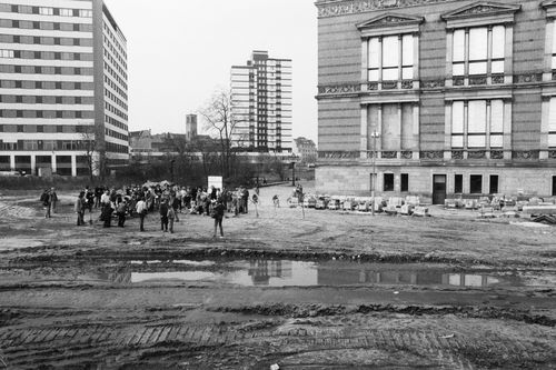 Eine Menschengruppe steht auf einem unbefestigten Gelände neben einem historischen Gebäude. Im Vordergrund eine große Pfütze und Reifenspuren, im Hintergrund zwei moderne mehrstöckige Gebäude.