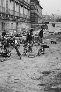 Fahrräder, ein Dreirad und umherstehende Menschen vor einem historischen Gebäude. Links verweist eine mehrsprachige Tafel auf die Zellen der Gestapo. Im Hintergrund die Berliner Mauer, ein Baugerüst mit Graffiti und mehrstöckige Gebäude.
