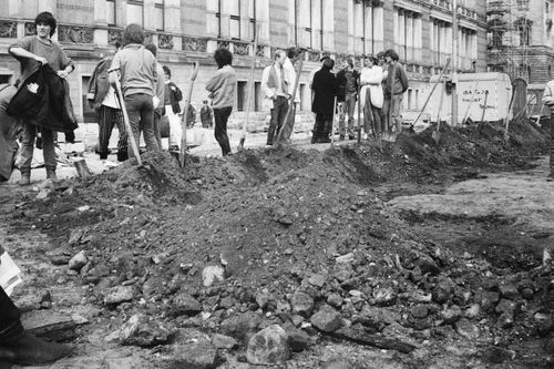 Ein Erdhaufen markiert den Verlauf eines Grabens, dessen lange Seite parallel zu einem historischen Gebäude verläuft. Die kurze Seite liegt im Vordergrund des Bildes. Entlang des Haufens stecken Spaten und Schaufeln, im Hintergrund stehen Menschen.