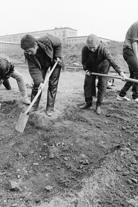 Eine Gruppe gräbt mit Spaten entlang eines rechtwinkligen, flachen Grabens. Die Grabenecke liegt unten im Vordergrund, mehrere Personen arbeiten gebeugt. Die Reihe setzt sich nach rechts und links fort.