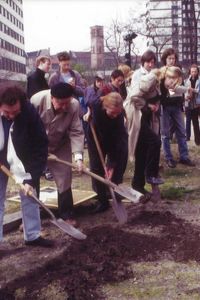 Sechs Personen graben mit Spaten entlang einer geraden Linie, eine siebte im rechten Winkel dazu. Der Graben ist flach, sie haben gerade begonnen. Menschen beobachten das Geschehen, einige halten Zettel.