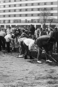 Viele Menschen graben mit Spaten auf einem unbefestigten Gelände. Sie arbeiten entlang einer Linie, die im rechten Winkel abknickt. Um sie herum stehen weitere Personen. Im Hintergrund moderne mehrstöckige Gebäude.