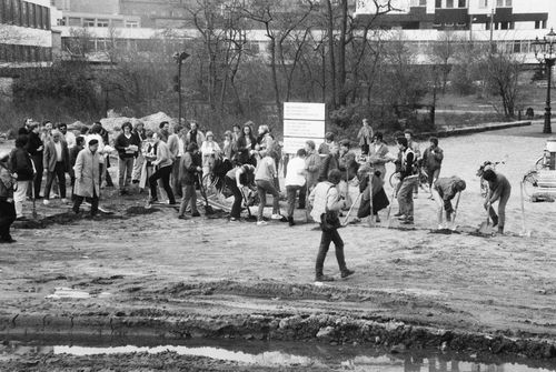 Zahlreiche Menschen versammeln sich auf einem unbefestigten Gelände. Einige stehen, andere graben mit Spaten. Im Vordergrund eine große Pfütze, im Hintergrund Bäume und moderne Gebäude.