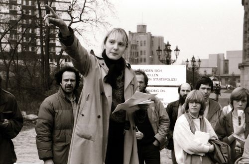 Eine Frau mit Zetteln in der Hand spricht und zeigt nach links. Umstehende Personen folgen der Geste mit dem Blick. Hinter ihnen ein Schild mit einem Hinweis auf ehemalige Gestapo-Folterzellen.