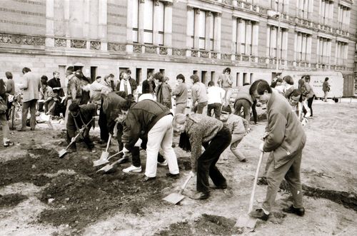Eine Gruppe von Menschen, dicht nebeneinander stehend, gräbt mit Spaten eine L-förmige Rinne in dunkler Erde vor einem historischen Gebäude. In der Mitte des rechtwinkligen Grabens steht ein Kind mit Spaten. Im Hintergrund ein Bauwagen und Zuschauende.