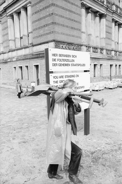 Eine Person trägt mehrere Spaten auf der Schulter und geht an einem Schild vorbei mit der Aufschrift: „Hier befanden sich die Folterzellen der geheimen Staatspolizei“, darunter Übersetzungen auf Englisch und einer weiteren Sprache.