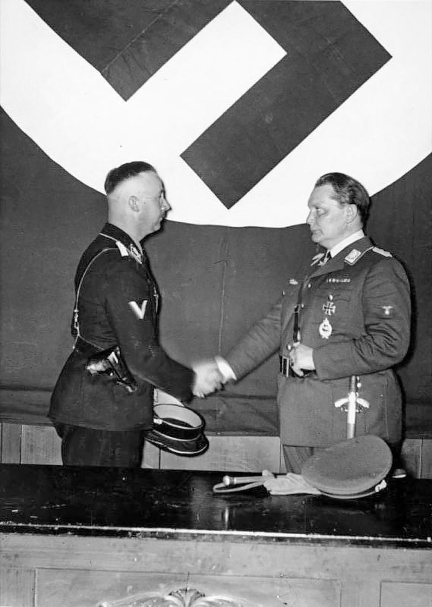 Schwarz-weiß-Fotografie: Zwei Männer in nationalsozialistischer Uniform schütteln sich mit ernstem Blick die Hände. Der Mann links trägt eine Pistole im Holster, der rechte einen Dolch. Sie stehen vor einer großen Hakenkreuzfahne an der Wand.