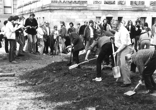 Im Vordergrund graben fünf Personen mit Spaten in dunkler Erde. Dahinter steht eine größere Gruppe dicht beieinander vor einem historischen Gebäude und beobachtet das Geschehen, einige mit Papieren oder Kameras.