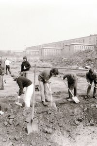 In einer Reihe graben Menschen mit Spaten auf einem offenen, steinigen Gelände eine rechtwinklige Rinne. In der Rinne steckt eine Schaufel senkrecht im Boden. Dahinter stehen einige Personen und beobachten. Im Hintergrund Hügel, Mauer und Gebäude.