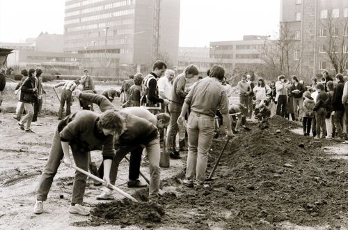 Menschen graben nebeneinander mit Spaten eine L-förmige Rinne. Entlang der Rinne türmt sich ausgehobene Erde. Einige beugen sich angestrengt, andere pausieren. Rechts viele Beobachtende, im Hintergrund zwei Personen mit Megafon.