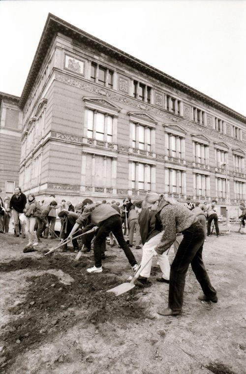 Mehrere Personen graben mit Spaten in dunkler Erde. Die Grabung hat gerade begonnen. Im Hintergrund ein mit Stuck verziertes, historisches Gebäude.