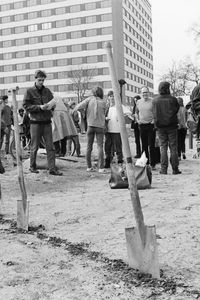 Entlang einer in die Erde gezogenen Linie stecken Spaten. Rechts liegt eine Schaufel auf dem Boden. Dahinter stehen mehrere Personen auf einem sandigen Gelände zwischen Wohnhäusern. Einige lesen von Zetteln oder unterhalten sich.