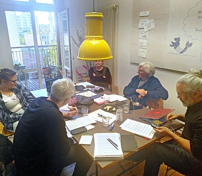 Sechs Personen sitzen um einen Tisch in einem hellen Raum. Auf dem Tisch liegen Notizen, Bücher und Snacks. An der Wand hängen Papiere mit Begriffen wie „Aktion“, „Ort“ und „Kontext“. Die Atmosphäre wirkt lebendig und gemeinschaftlich.
