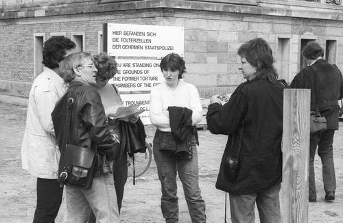 Vor einem mehrsprachigen Schild zu ehemaligen Folterzellen der geheimen Staatspolizei stehen sechs Personen und scheinen sich über den historischen Ort auszutauschen.