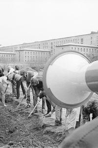 Rechts im Vordergrund eine Person mit Brille, die in ein Megafon spricht. Links im Bildrand sind Textblätter zu sehen. Eine lange Reihe Menschen gräbt konzentriert einen schmalen Graben. Im Hintergrund ein massives Gebäude mit klar gegliederter Fassade.