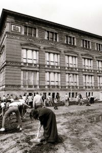 Vor einem historischen Gebäude mit stuckverzierter Fassade graben Menschen mit Spaten eine rechtwinklige Rinne. Rechts im Bild große Pfützen und deutliche Reifenspuren im unebenen Boden.