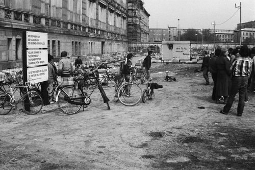 Fahrräder, ein Dreirad und umherstehende Menschen vor einem historischen Gebäude. Links verweist eine mehrsprachige Tafel auf die Zellen der Gestapo. Im Hintergrund die Berliner Mauer, ein Baugerüst mit Graffiti und mehrstöckige Gebäude.
