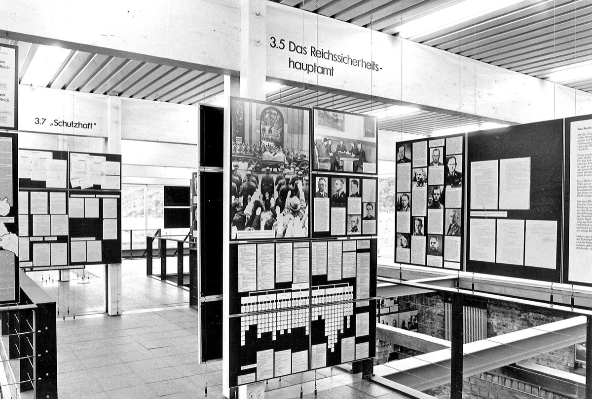 Schwarz-weiß-Fotografie: Blick in eine Ausstellung mit zahlreichen frei hängenden Text- und Bildtafeln.