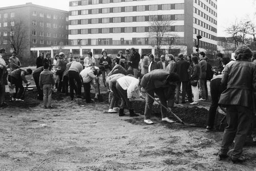 Viele Menschen graben mit Spaten auf einem unbefestigten Gelände. Sie arbeiten entlang einer Linie, die im rechten Winkel abknickt. Um sie herum stehen weitere Personen. Im Hintergrund moderne mehrstöckige Gebäude.