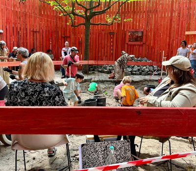 Zwei Frauen sitzen im Vordergrund auf einer roten Bank und beobachten eine Gruppe von Menschen – darunter Kinder – beim Graben in einem sandigen Bereich. Dieser ist von roten Holzwänden umgeben.