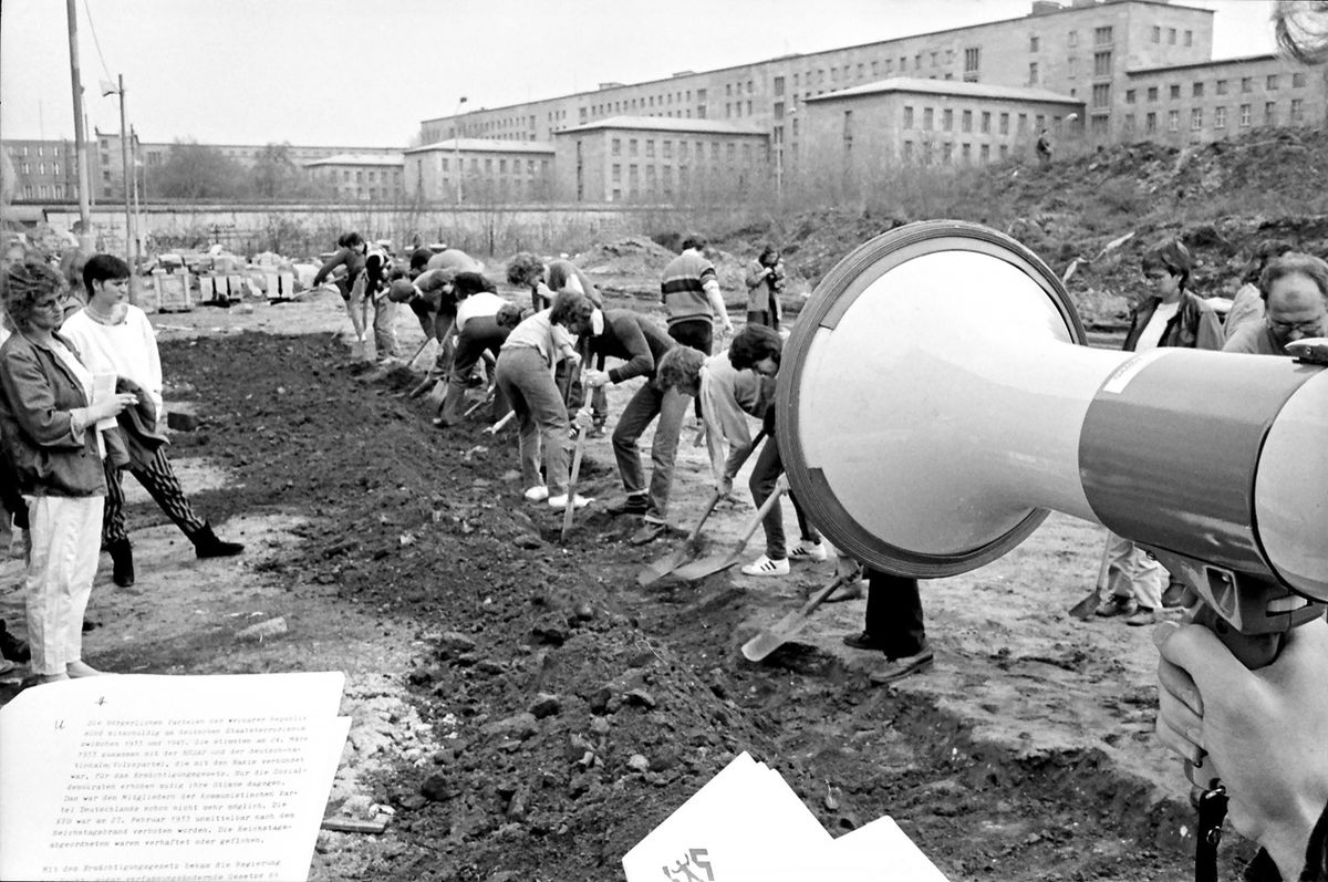 Schwarz-weiß-Fotografie: Eine lange Reihe Menschen gräbt konzentriert einen schmalen Graben. Rechts im Vordergrund ein Megafon und Textblätter in einer Hand. Im Hintergrund ein massives Gebäude mit klar gegliederter Fassade.