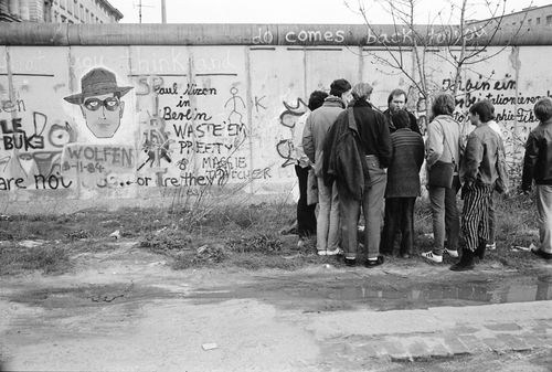 Eine kleine Gruppe von Personen steht dicht beisammen vor der Berliner Mauer, die mit zahlreichen Graffiti besprüht ist. Das Graffiti zeigt unterschiedliche Schriftzüge und ein Gesicht mit Hut und Brille. Vor der Mauer eine Wasserpfütze und niedriges Gras