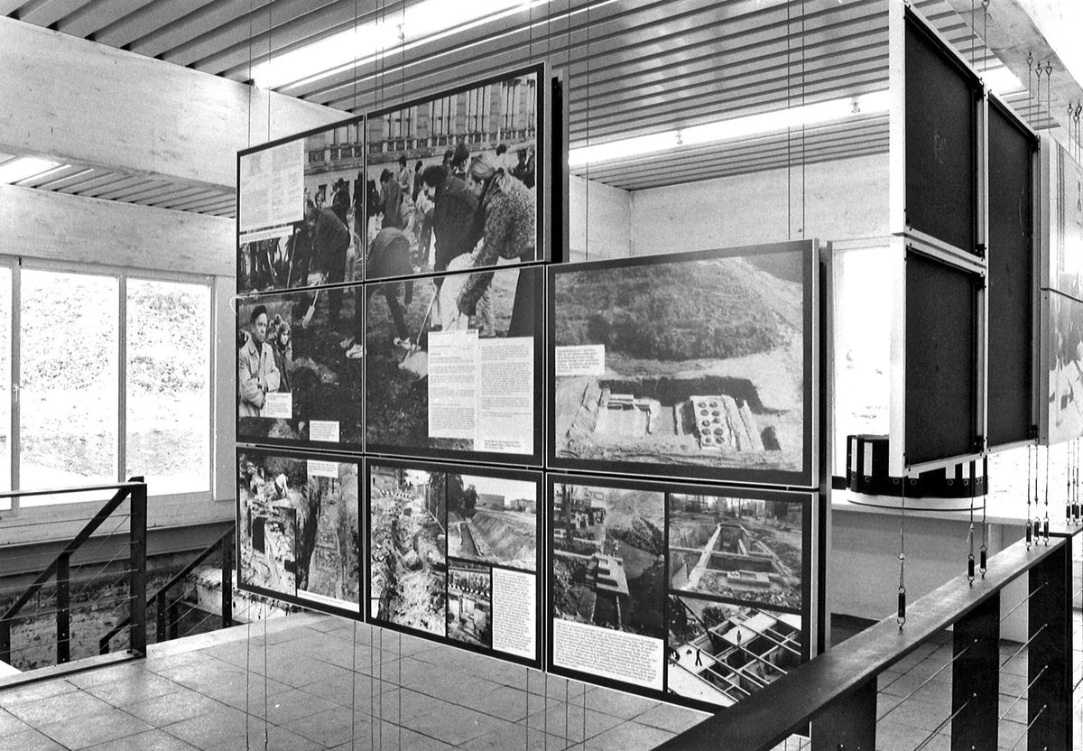 Schwarz-weiß-Fotografie: Blick in eine Ausstellung mit großformatigen Informationstafeln, die frei im Raum hängen. Die Tafeln zeigen Texte und Bilder zur Grabungsdokumentation.