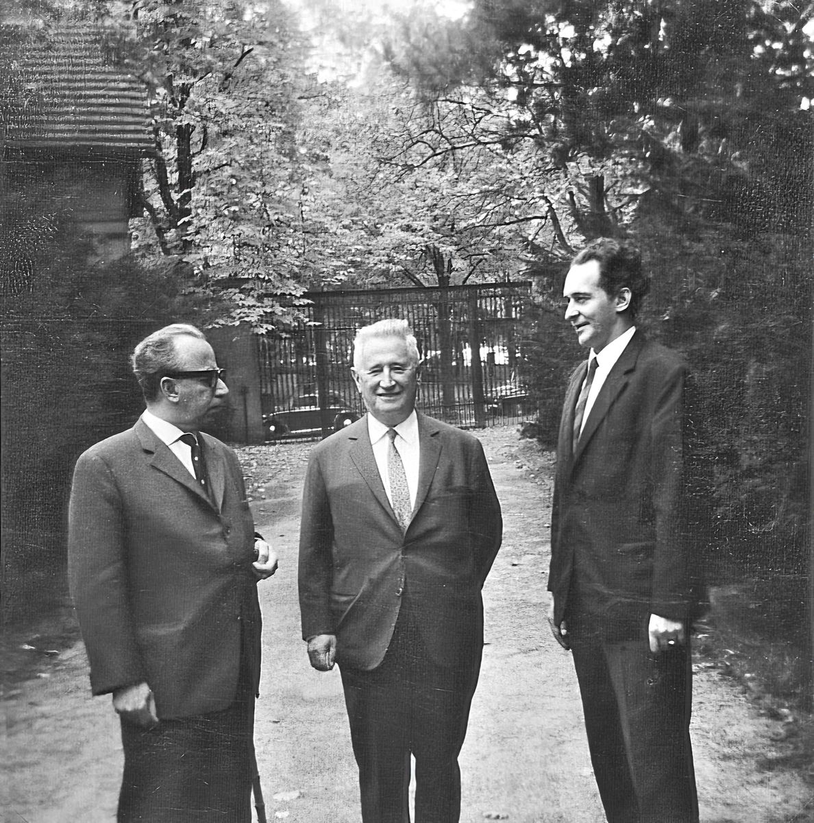 Schwarz-weiß Fotografie von drei Männer im Anzug im Freien; der mittlere lächelt in die Kamera, der rechte blickt freundlich zum linken, der ernst schaut.