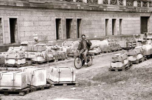 Ein Mann, der mit einem Fahrrad durch eine Baustelle mit gestapelten Steinquadern fährt. Mit einer Hand geschultert trägt er einen Spaten. Im Hintergrund ein historisches Gebäude mit Stuckverzierter Fassade.