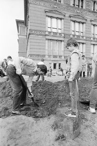 Ein Mann beugt sich mit einem Spaten über aufgewühlte Erde. Rechts daneben steht ein Kind auf einem kleinen Steinquader und schaut hinab. Im Hintergrund ein großes historisches Gebäude mit verzierter Fassade und weiteren Personen.