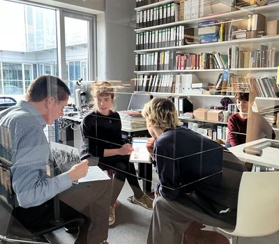 Vier Personen sitzen in einem hellen Büroraum an einem Tisch, gesehen durch eine Glasscheibe. Die Wände sind mit Regalen voller Bücher und Aktenordner gesäumt. Sie besprechen Dokumente, die Atmosphäre wirkt konzentriert und gemeinschaftlich.