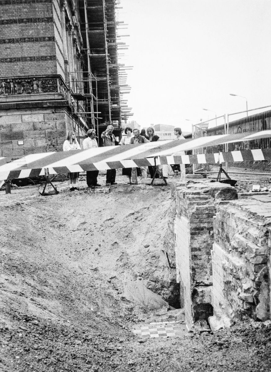 Schwarz-weiß-Fotografie: Eine Gruppe Menschen blickt hinter einer Absperrung in eine Ausgrabung mit freigelegten Mauerresten und einem gefliesten Bodenstück. Links ein historisches Gebäude mit Baugerüst, rechts im Hintergrund die Berliner Mauer.