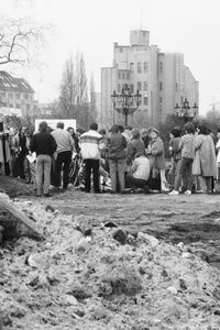 Eine große Menschengruppe steht auf einem unbefestigten Gelände neben einem historischen Gebäude. Im Vordergrund ein Hügel aus aufgeworfener Erde und Reifenspuren, im Hintergrund Bäume und mehrstöckige Gebäude.