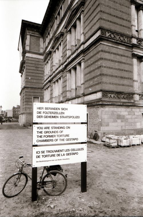 Ein Schild vor einer historischen Gebäudeecke trägt den Hinweis: „Hier befanden sich die Folterzellen der Geheimen Staatspolizei.“ Darunter Übersetzungen auf Englisch und Französisch. Links lehnt ein Fahrrad am Schild.