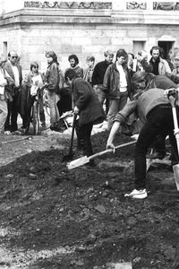 Im Vordergrund graben fünf Personen mit Spaten in dunkler Erde. Dahinter steht eine größere Gruppe dicht beieinander vor einem historischen Gebäude und beobachtet das Geschehen, einige mit Papieren oder Kameras.